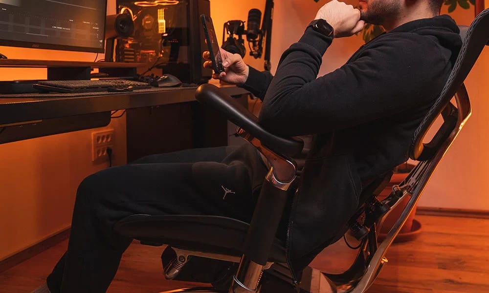 Un homme assis sur une chaise ergonomique regardant son téléphone, le dossier épouse la courbure du dos et des cervicales pour un bon soutien.