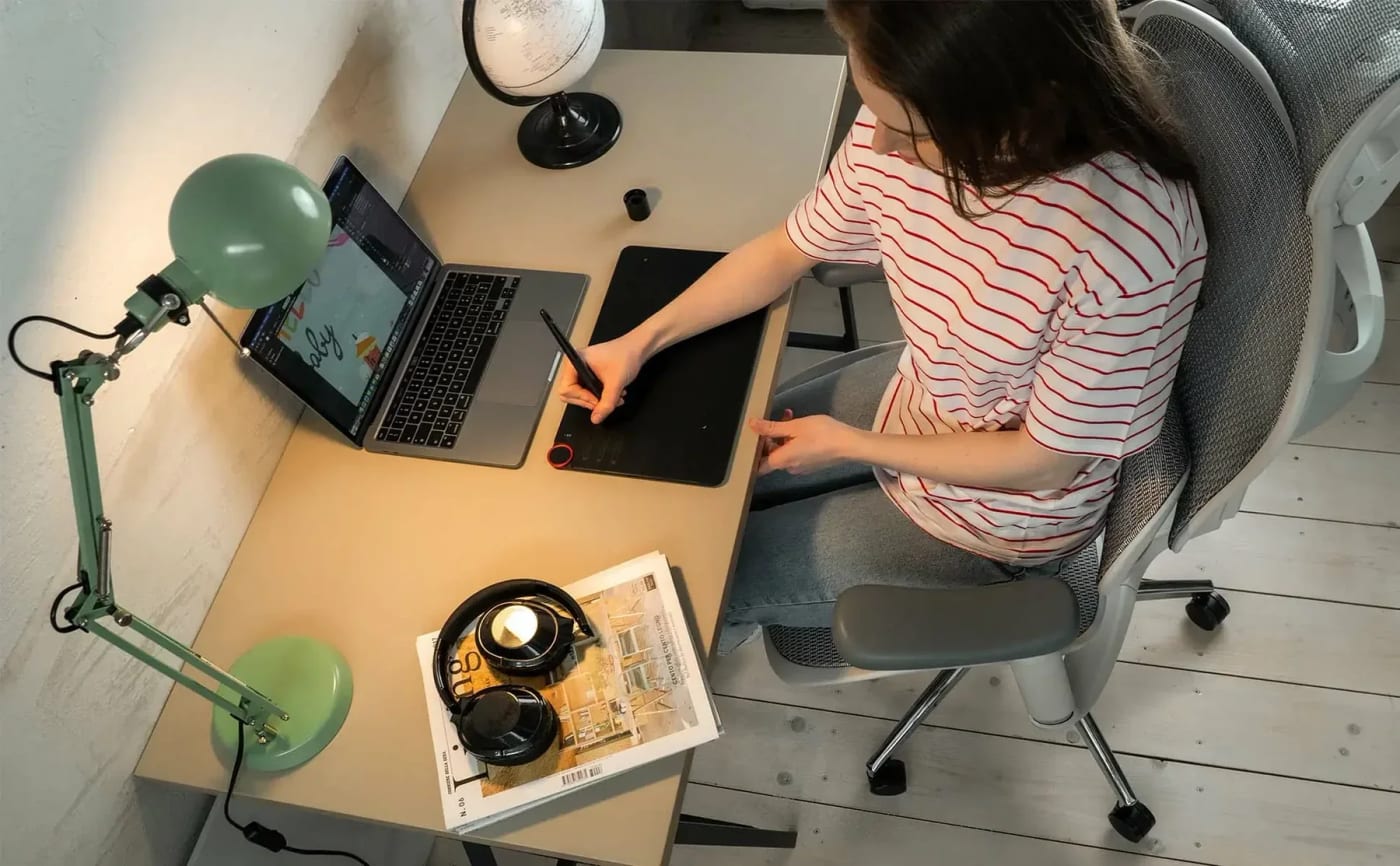 Une femme vêtue d’un t-shirt rayé rouge et blanc est assise sur une chaise ergonomique, utilisant une tablette numérique pour créer sur un ordinateur portable, avec une lampe de bureau verte, un globe, des magazines et des écouteurs sur la table.